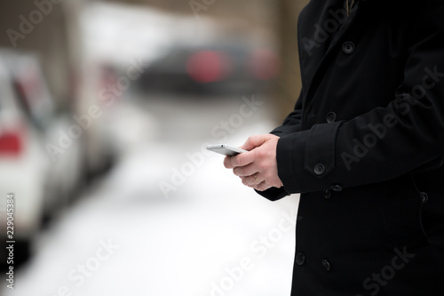 Businessman holding a smartphone mobile, stuck in traffic on a snowy cold winter day. Man in coat texting, cars in the background.