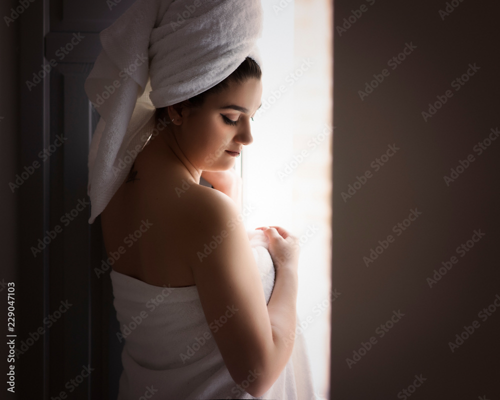 Fototapeta premium Portrait of a beauty girl with a towel in her head and a towel around her body looking through the window