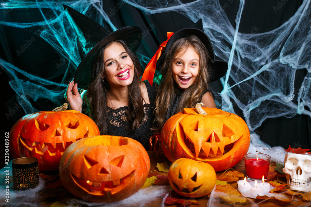 Fototapeta premium Happy halloween. Cute little girl and her beautiful mom in witch hats are looking at camera and smiling while sitting on wooden background decorated for Halloween