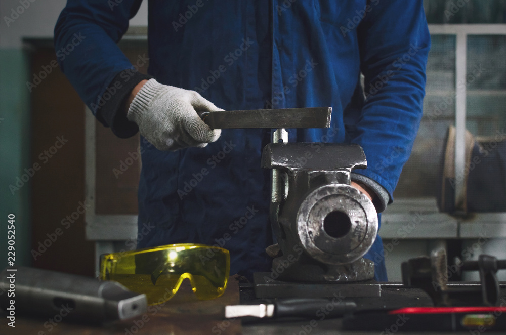 Man worker is working with iron detail on vise grip on blacksmith table and using a file.