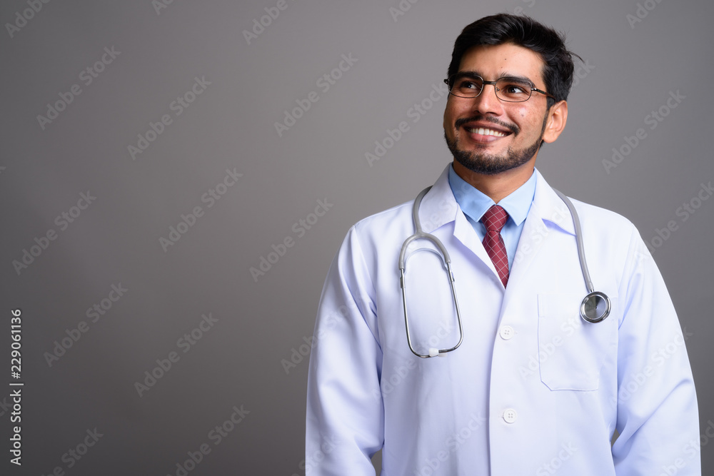 Young handsome bearded Persian man doctor against gray backgroun Young handsome bearded Persian man doctor against gray backgroun
