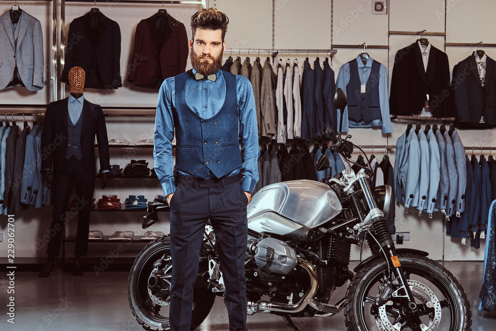 Elegantly dressed man posing with hands in pockets near retro sports motorbike at the men's clothing store.