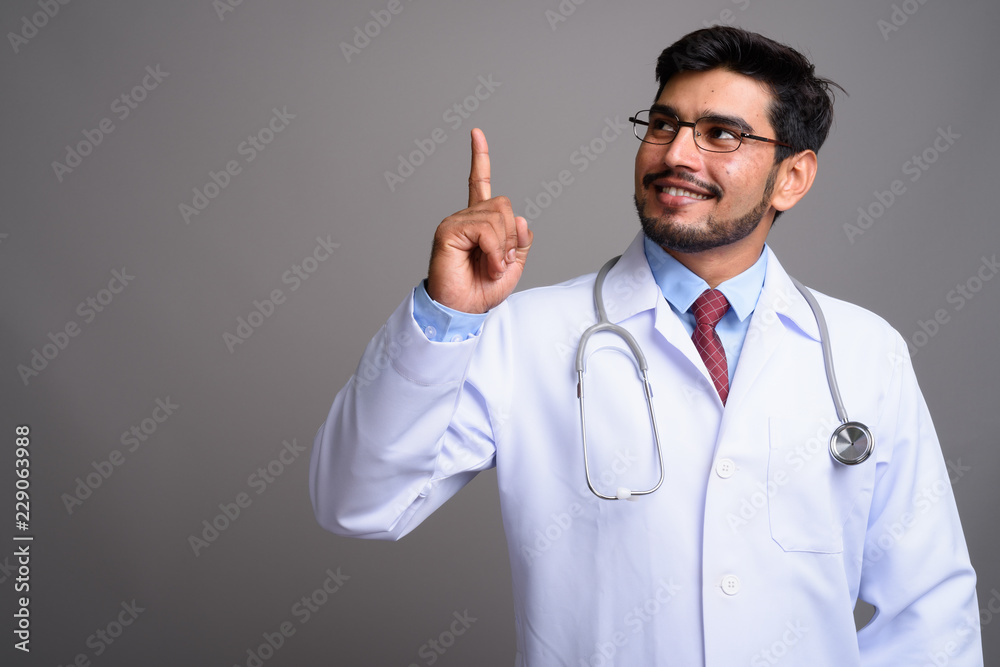 Young handsome bearded Persian man doctor against gray backgroun Young handsome bearded Persian man doctor against gray backgroun