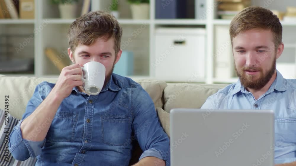 Medium tracking shot of two young twin brothers sitting on sofa in living room at home and talking to each other, one of them drinking tea or coffee and one using laptop computer Medium tracking shot of two young twin brothers sitting on sofa in living room at home and talking to each other, one of them drinking tea or coffee and one using laptop computer
