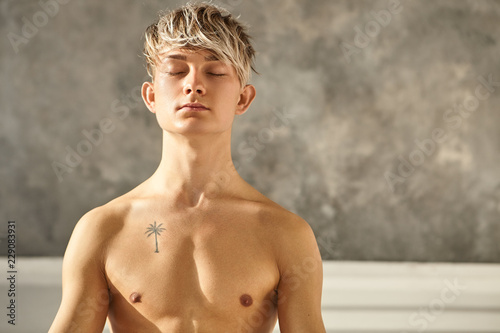 Portrait of handsome tattooed man practicing yoga indoors, closing eyes while meditating, having peaceful look, concentrating on his breath. Shirtless young male teacher doing meditaiton at gym photo