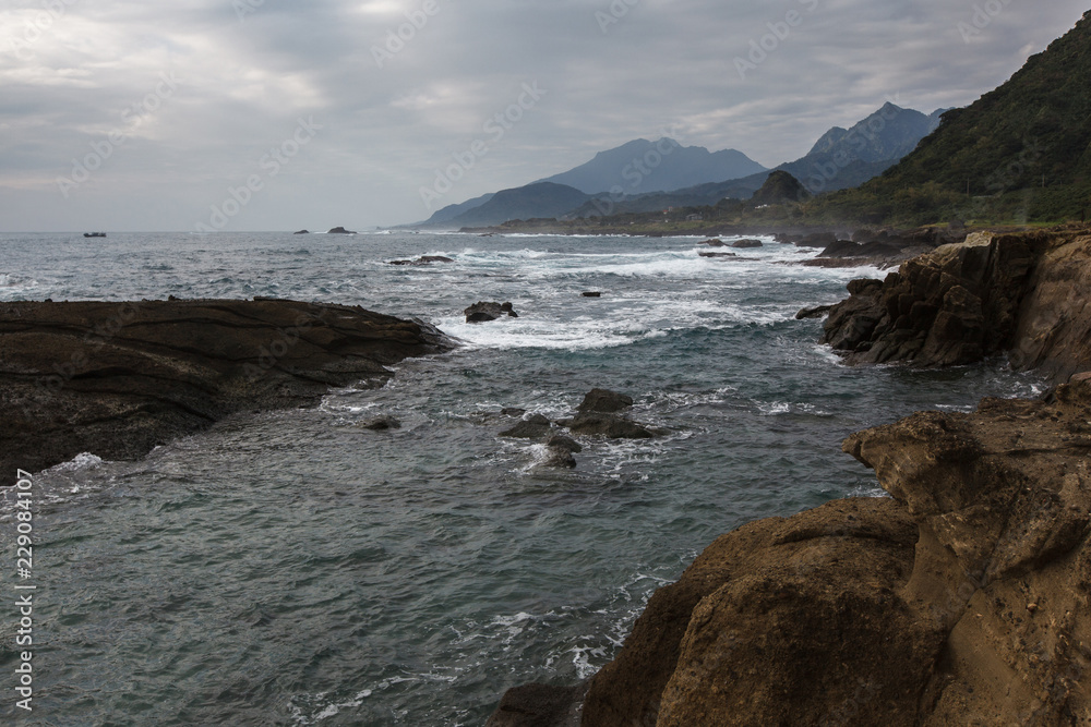 Taiwan East Coast Rocky Coastline Background Image - Overcast Skies, Exotic Rock Formations, Grass and Waves in the Ocean. Ocean Coastline, Asia Landscape Photography, Rays of sunshine in background