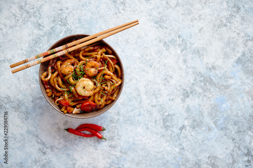 Traditional asian udon stir-fry noodles with shrimp in bowl and chopsticks. Fresh chilli pepers on side. Placed on bright stone background with copy space.