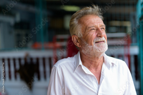 Face of happy senior bearded man smiling while thinking outdoors