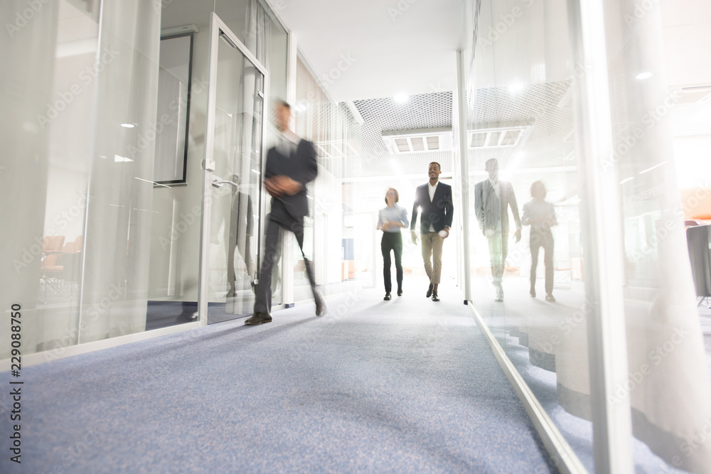 Blurred motion of modern business people crossing illuminated office corridor while hurrying to their workplaces, corporate company