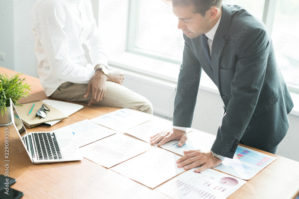 Concentrated young businessman in formal suit leaning on table with financial papers and analyzing documents while preparing for annual meeting Concentrated young businessman in formal suit leaning on table with financial papers and analyzing documents while preparing for annual meeting