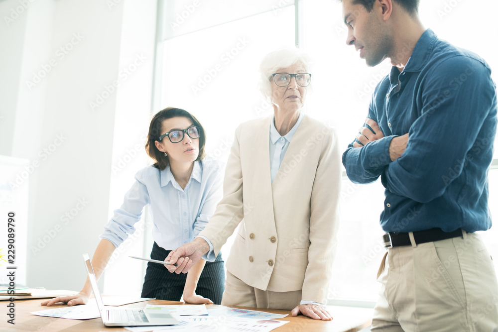 Serious confident senior lady in stylish jacket pointing at laptop screen while explaining analytical data to colleagues at meeting in office Serious confident senior lady in stylish jacket pointing at laptop screen while explaining analytical data to colleagues at meeting in office