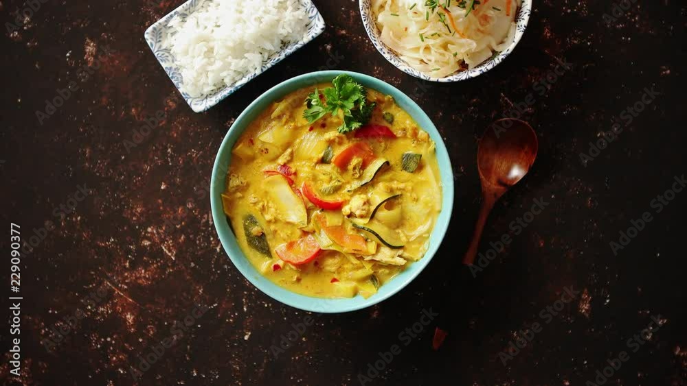 Traditional Chinese or Thai chicken yellow curry with vegetables served with steamed rice cucomber and hot chilli pepper on side. Top view with copy space on dark rusty background.