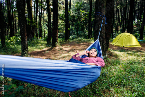 Smiling asian hiker woman talking on cellphone while lying in hammock