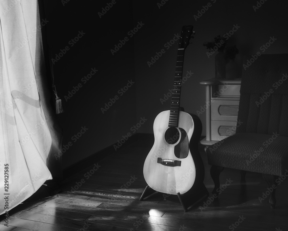 Fototapeta Black & white photo of acoustic Spanish guitar on a stand in the moody shadows of a dark room with bright light coming in from behind a curtain