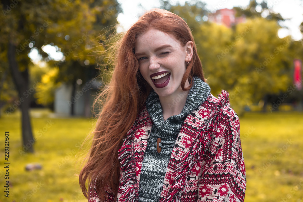 Redhead young happy cheerful woman with freckles and dark lipstick posing outdoors in park. Looking camera winking.