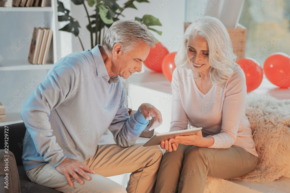 Obraz premium Valuable memories. Charming elderly woman siting in living room with a tablet and showing pictures to her pleased positive husband sitting next to her