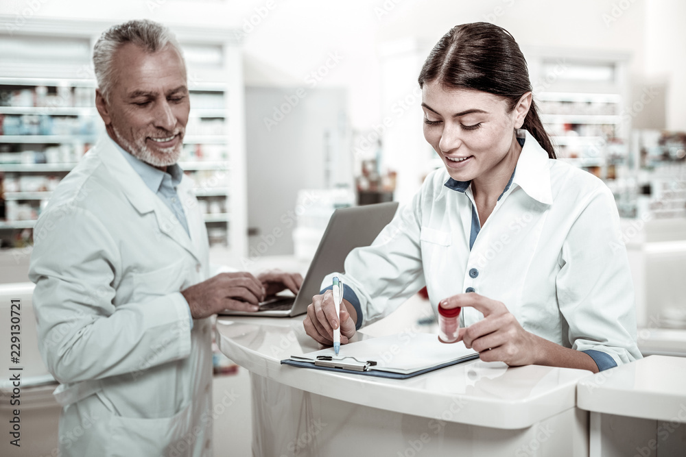 Work in drugstore. Dark-haired beaming experienced woman working in drugstore feeling busy receiving new medicine Work in drugstore. Dark-haired beaming experienced woman working in drugstore feeling busy receiving new medicine
