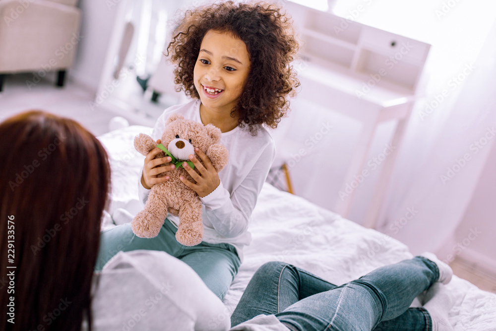 My toys. Joyful child sitting on bed and looking at her young mother My toys. Joyful child sitting on bed and looking at her young mother