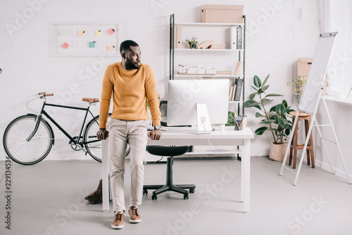 handsome african american designer in orange sweater leaning on table and looking away in office