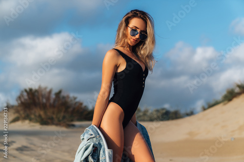 Sexy girl on the beach in a black swimsuit and blue sunglasses with a denim jacket in her hands. Beautiful model with a sports figure walks near the ocean on vacation, enjoying nature.