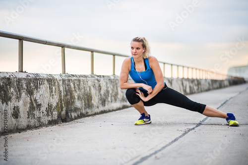 Young sporty woman runner with earphones stretching by the sea outside. © Halfpoint