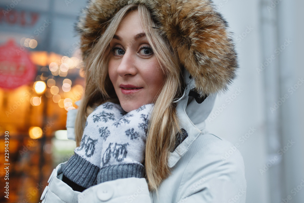 Attractive, charming smiling young woman in a white coat with fur hood walking in the winter street and warms frozen hands in white mittens. Background blurry lights. The holiday mood. Attractive, charming smiling young woman in a white coat with fur hood walking in the winter street and warms frozen hands in white mittens. Background blurry lights. The holiday mood.