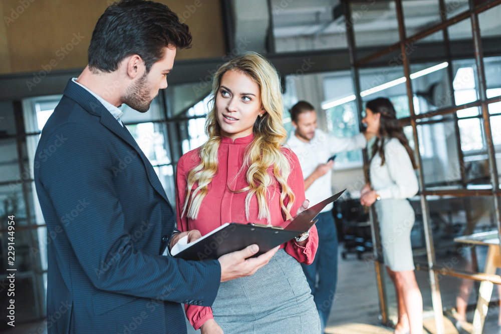 businesspeople discussing work while colleagues having conversation behind in office businesspeople discussing work while colleagues having conversation behind in office