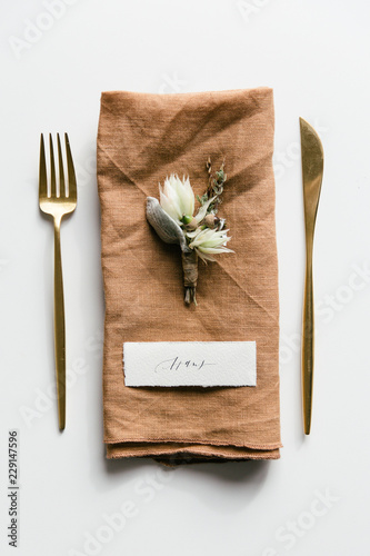 Table setting with golden colored cutlery styled with a orange linnen napkin, boutonniere and handwritten place card.