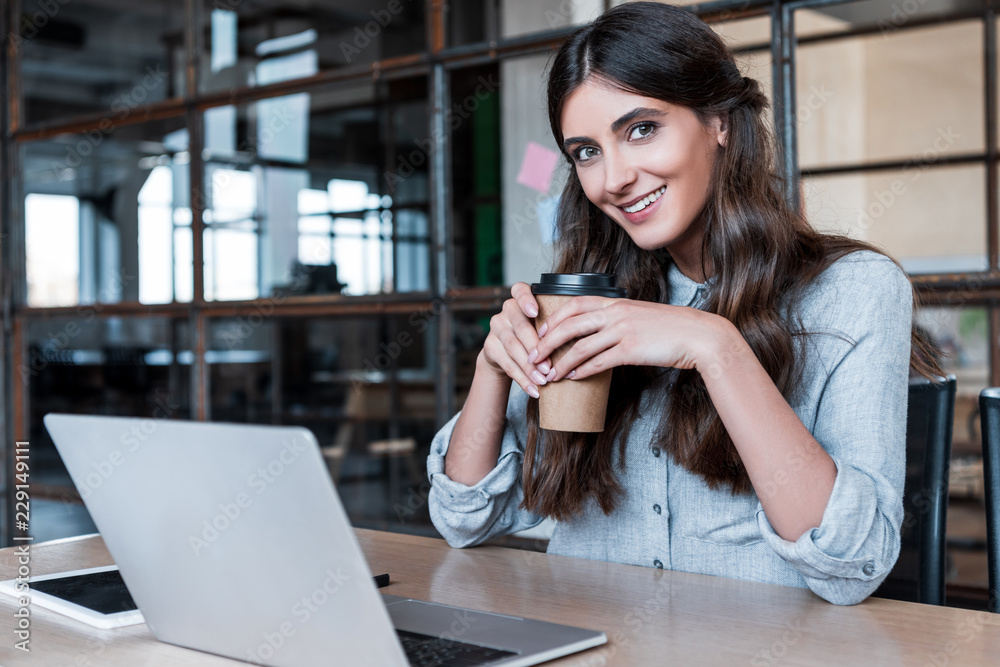 happy young businesswoman holding paper cup and smiling at camera while working with laptop in office