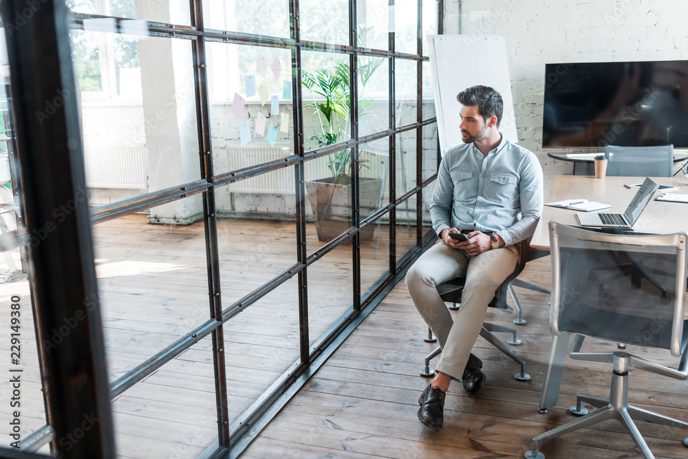 Fototapeta premium high angle view of young businessman using smartphone and looking away while sitting in office