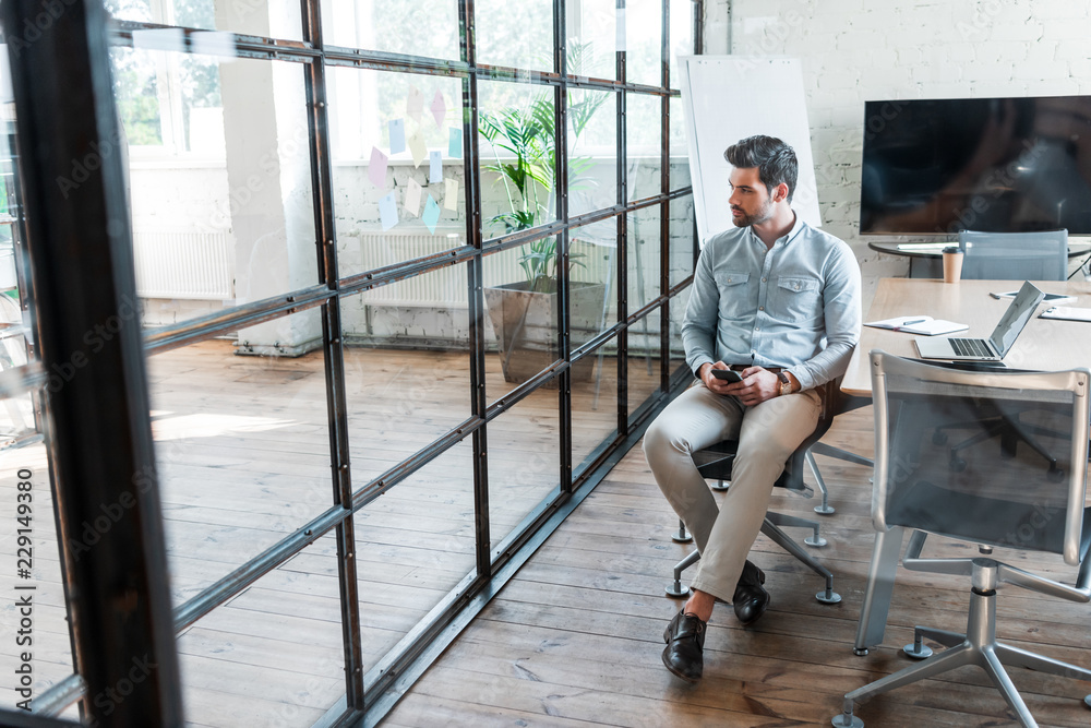 high angle view of young businessman using smartphone and looking away while sitting in office