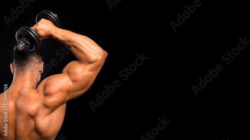 Fit muscular man uses his dumbbell to work his triceps on dark background
