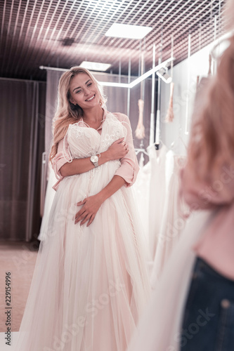 Perfect mood. Joyful young woman smiling to her reflection while holding a wedding dress