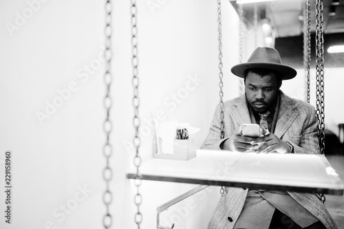 Stylish African American man model in gray coat, jacket tie and red hat sitting on swing at cafe with mobile phone at hands. Black and white photo. photo