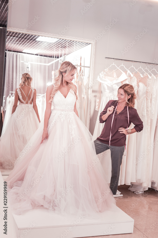 Professional service. Nice female tailor holding her glasses while looking at the bride Professional service. Nice female tailor holding her glasses while looking at the bride