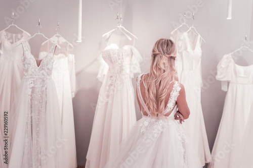 Difficult choice. Beautiful blonde woman standing in front of wedding dresses while thinking about what to choose