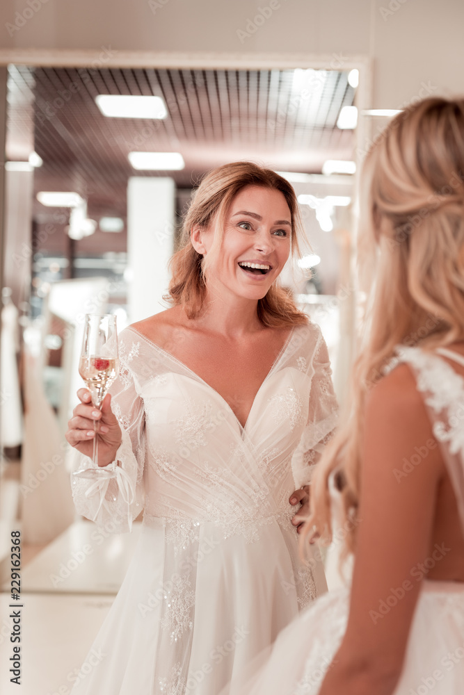 Pleasant interaction. Happy joyful woman standing with a glass of champagne while listening to her friend Pleasant interaction. Happy joyful woman standing with a glass of champagne while listening to her friend