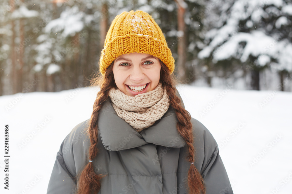 Cheerful excited attractive student girl in knitted hat and scarf laughing while looking at camera, she walking in winter forest Cheerful excited attractive student girl in knitted hat and scarf laughing while looking at camera, she walking in winter forest