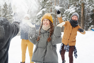 Happy young friends in winter coats having snowball fight in forest: excited girl standing in center and feeling snow on face