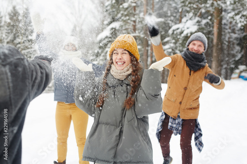 Happy young friends in winter coats having snowball fight in forest: excited girl standing in center and feeling snow on face