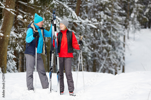 Active senior spouses having talk in winter forest while moving down snowdrift for skiing training
