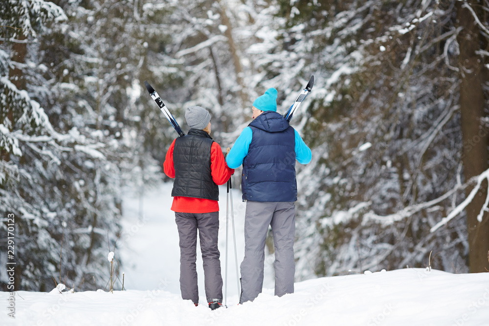 Rear view of sporty aged couple with skiing equipment walking down forest path on winter day