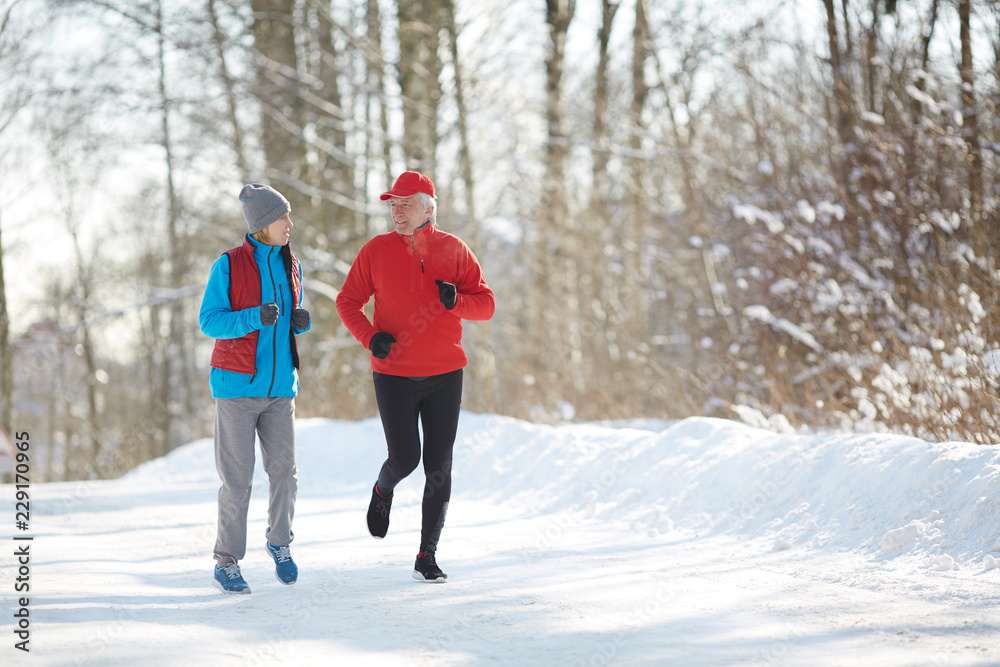 Active senior couple in sportswear running down snow road in winter forest on frosty morning