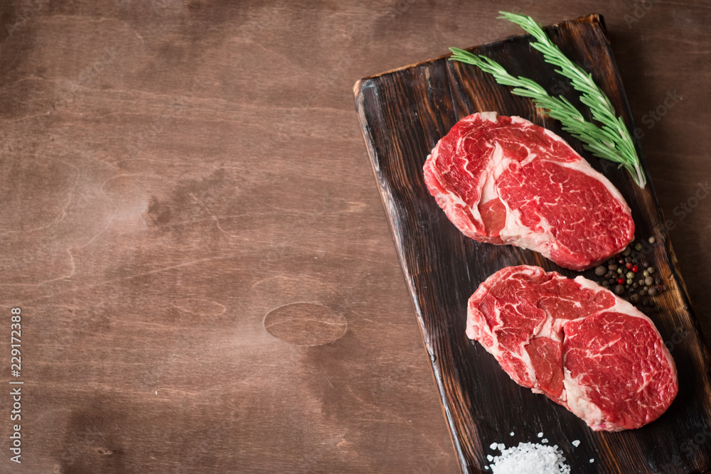 Two fresh raw rib-eye steak on wooden Board on wooden background with salt, pepper and rosmary in a rustic style, top view