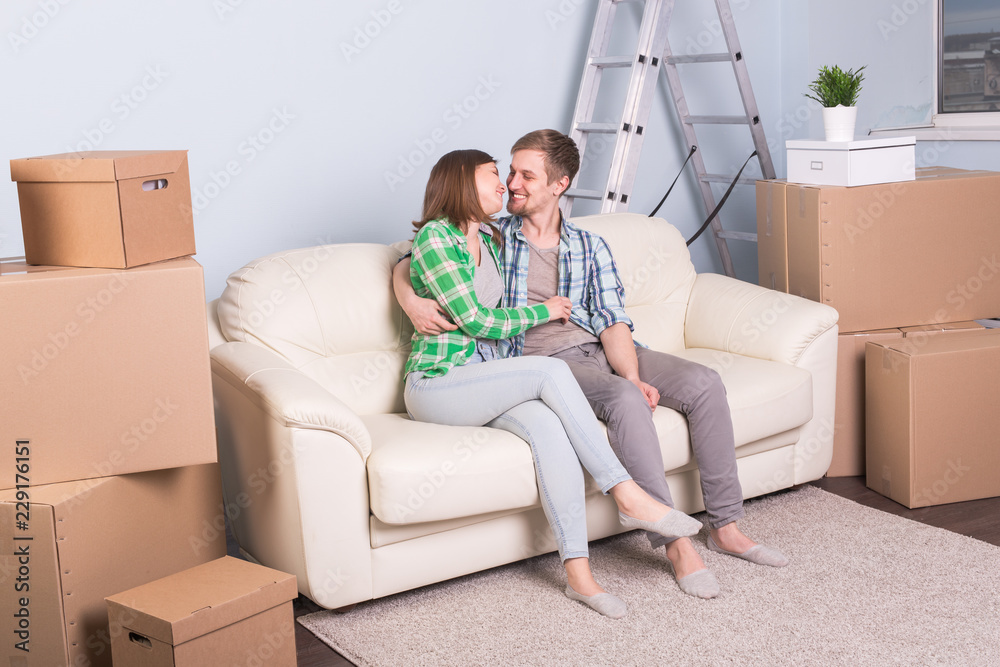 Moving, new apartment and relocation concept - young couple sitting and hugging on the couch surrounded with boxes