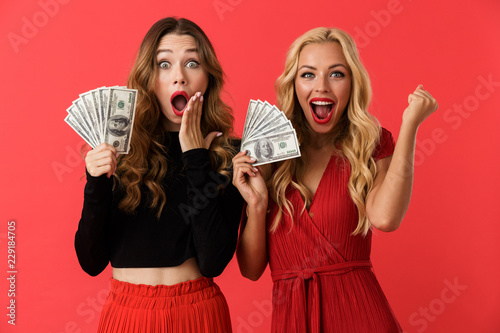 Emotional excited young friends women standing isolated over red background holding money.
