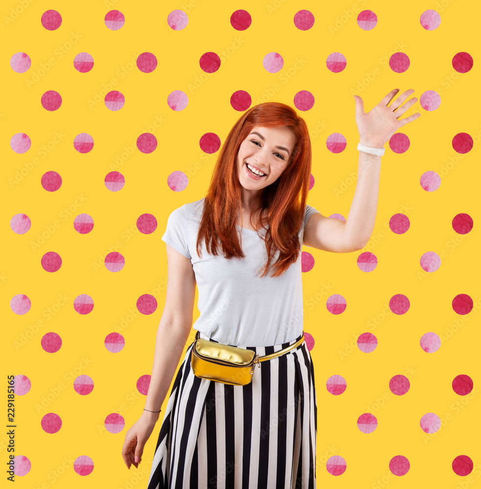 Hello. Cheerful young girl smiling and waving her hand while standing against the bright dotted background Hello. Cheerful young girl smiling and waving her hand while standing against the bright dotted background