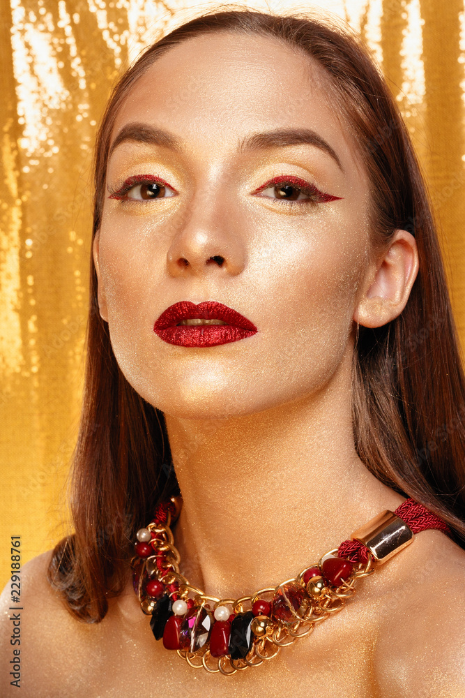 Magic Girl Portrait in Gold. Golden Makeup, close-up portrait in studio  shot, color. Beauty Model Girl with perfect bright make-up, red lips,  golden maroon jewellery. Sexy lady makeup Holiday party. Stock Photo