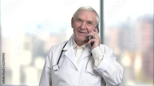 Handsome senior old doctor in white coat and stethoscope is talking on the mobile phone in hospital. Bright abstracrt blurred windows background with view on city. photo