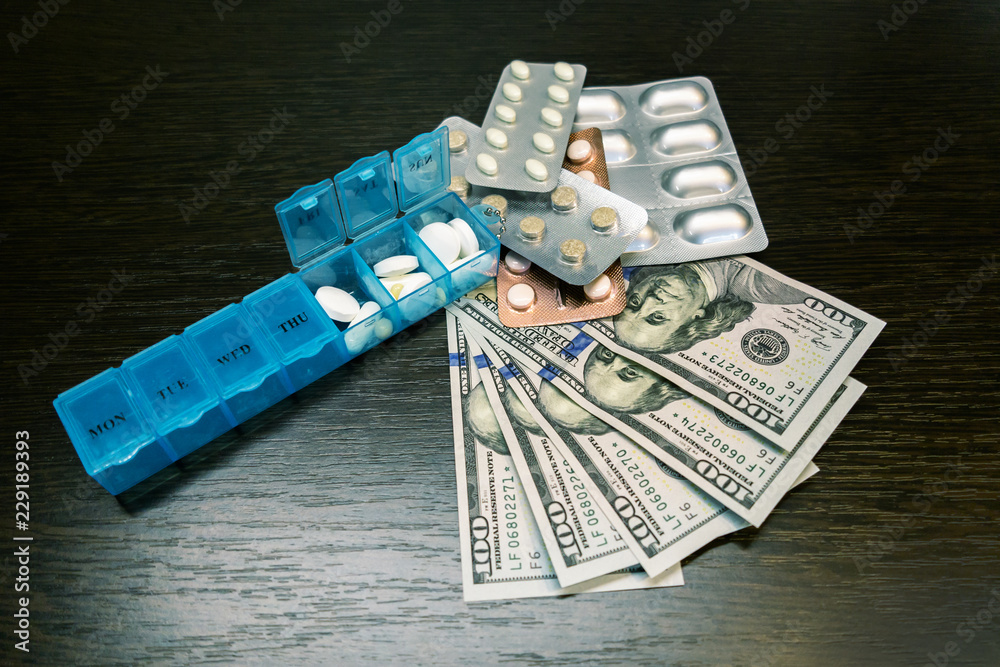 Pillbox, pills and tablets on dollar money on dark wooden table. Medicine expenses. High costs of medication concept Pillbox, pills and tablets on dollar money on dark wooden table. Medicine expenses. High costs of medication concept
